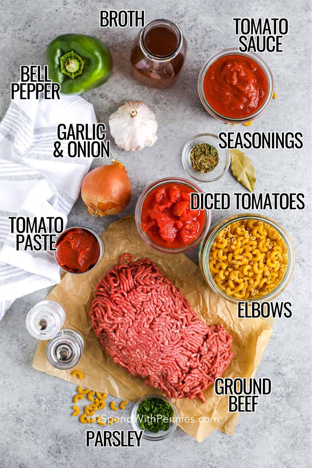 ingredients for goulash on a table including broth, tomato sauce, bell pepper, seasonings, garlic, onion, diced tomatoes, tomato paste, pasta, and ground beef 